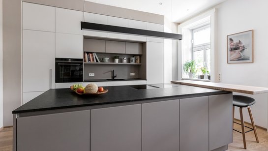 Hellgraue Kücheninsel mit seitlicher Barplatte und Barhockern, dahinter weiße Hochschränke mit einer Nische