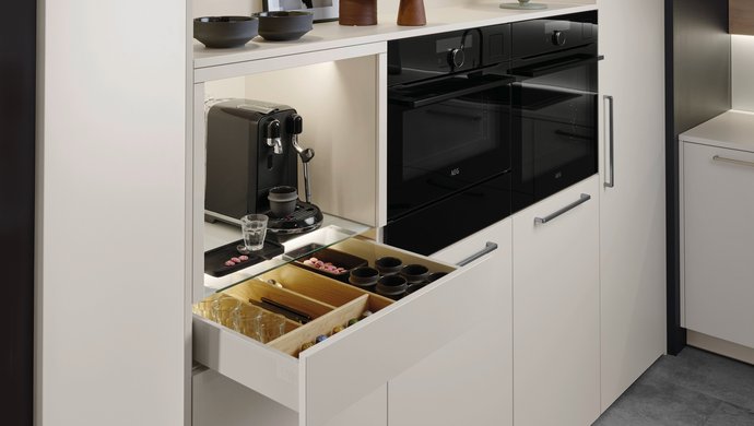 Helle Hochschrankgruppe mit einer Nische für Kaffeemaschinen, die Lade darunter für Kaffeezubehör ist geöffnet
