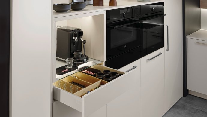 ewe Nuova/Cemento in angora/patina bronze Helle Hochschrankgruppe mit einer Nische für Kaffeemaschinen, die Lade darunter für Kaffeezubehör ist geöffnet