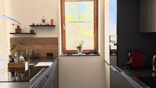 Blick zwischen Kochinsel und Küchenzeile: graue Auszüge links, helle und graue Küchenschränke mit eingebautem Backofen und Spülbereich rechts, Fenster am Ende des Durchgangs.