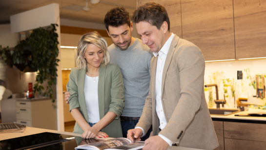 Persönliche und kompetente Beratung durch den ewe Küchenfachhändler Ein Küchenfachhändler in einem beigefarbenen Sakko berät ein Paar bei der Küchenplanung
