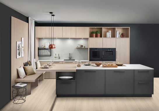 Große dunkle Kücheninsel vor einer Küchenzeile aus hellem Holz, links an der Insel ist eine Sitzbank