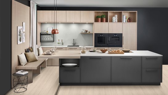 Große dunkle Kücheninsel vor einer Küchenzeile aus hellem Holz, links an der Insel ist eine Sitzbank