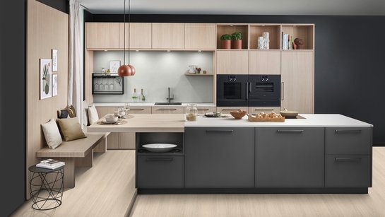 Große dunkle Kücheninsel vor einer Küchenzeile aus hellem Holz, links an der Insel ist eine Sitzbank