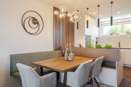 Offene Wohnküche mit Holzesstisch, gepolsterter Sitzbank und hellen Stühlen; ein Kräuterkasten mit Nussfurnier gliedert den Übergang zum Kochbereich.