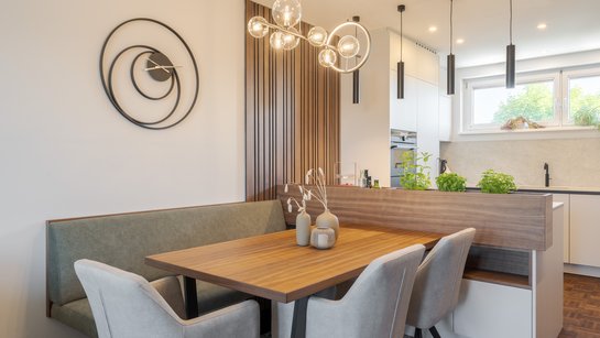 Offene Wohnküche mit Holzesstisch, gepolsterter Sitzbank und hellen Stühlen; ein Kräuterkasten mit Nussfurnier gliedert den Übergang zum Kochbereich.