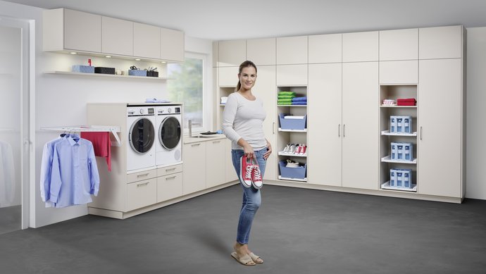 Eine Frau steht vor einem L-förmigen, großen Hauswirtschaftsraum mit weißen Fronten