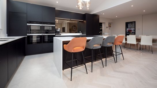 Kücheninsel mit seitlich herabgezogener Marmoroptik-Arbeitsplatte und Bartsühlen, dahinter eine schwarze Küche