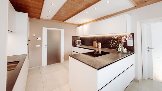 Moderne grifflose Küche in Steinweiß mit schwarzen Griffmulden, Naturstein-Arbeitsplatte und Rückwand, Holzdecke in Esche und weiß mit Spots.