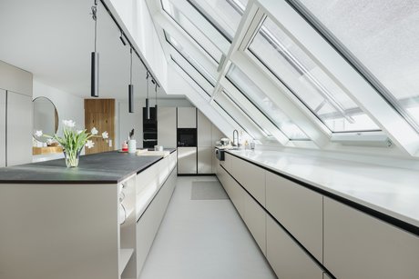 Blick durch eine helle Inselküche mit schwarzen Griffmulden unter einer schrägen Fensterfront