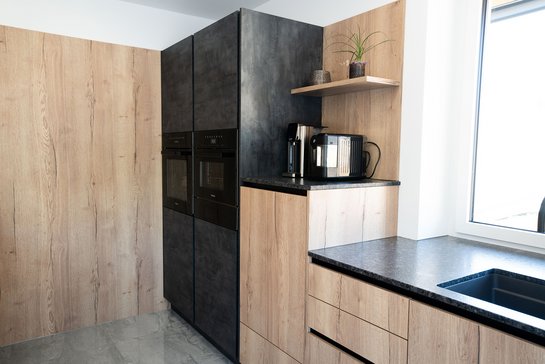Küchenzeile in Holzoptik mit dunkler Steinarbeitsplatte, dahinter ein Hochschrank in metallgrau