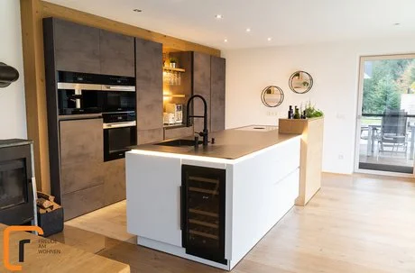 Eine weiße Kücheninsel mit schwarzer Arbeitsplatte, dahinter eine Küchenzeile in Betonoptik