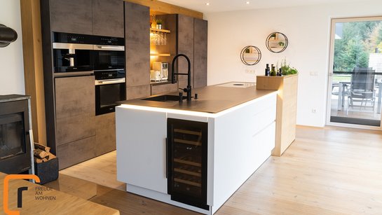 Eine weiße Kücheninsel mit schwarzer Arbeitsplatte, dahinter eine Küchenzeile in Betonoptik