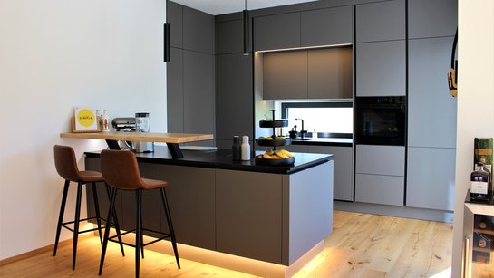 Frontalansicht der basaltgrauen ewe nanoo® mit Barbereich, zwei Hockern, LED-Beleuchtung und deckenhohen Hochschränken.