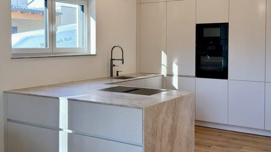 Kompakte U-Küche in angoraweiß mit Kochinsel, integriertem Kochfeld und hochgestellt eingebautem Backofen in klarer Linienführung.