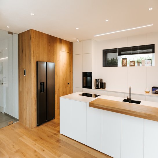 Weiße Kücheninsel mit aufgesetzter Thekenplatte aus Holz, links davon Hochschränke mit Holzfront.