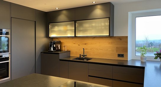 Ecke einer grauen Küche mit Hochschränken, Glas-Oberschränken und Rückwand aus Holz