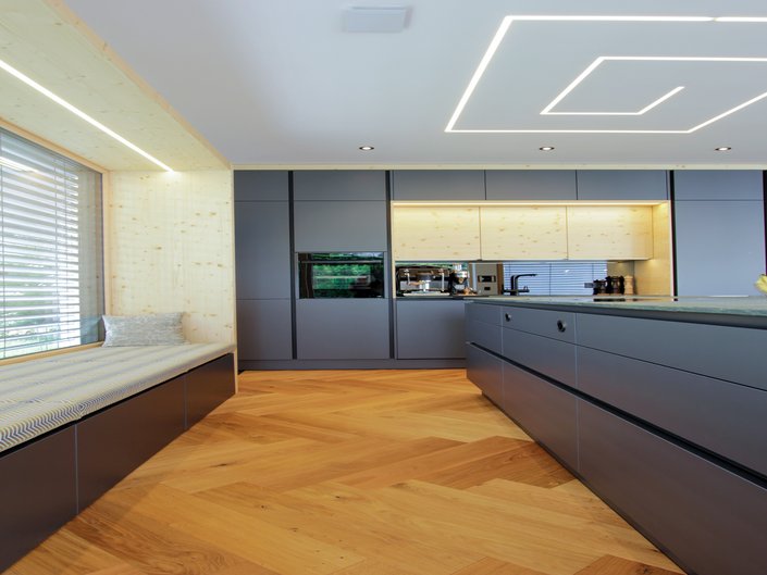 Rechts eine Kücheninsel und dahinter eine Küchenzeile in grau metallic, links eine Fenstersitzbank mit passenden Schubladenfronten