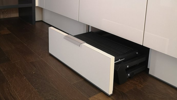 Aus einer weißen Küche wird eine Sockellade gezogen, in der sich eine Leiter befindet