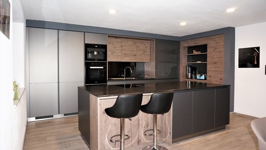 Inselküche mit besonders reinigungsfreundlicher Küchenoberfläche ewe nanoo® im Farbton carbon metallic kombiniert mit Holzelementen