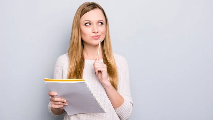 Eine junge blonde Frau hat einen Block und einen Stift in der Hand und überlegt intensiv.