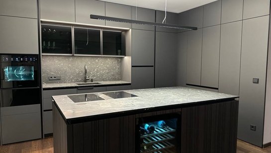 Dunkle Küche mit Kochinsel, strukturierten Fronten in Holzoptik, Stein-Arbeitsplatte und integriertem Weinkühlschrank vor raumhohen Schränken.
