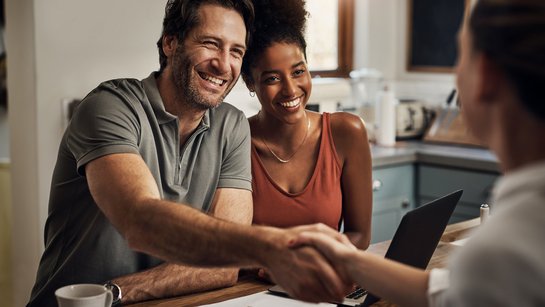 Junges Paar sitzt lächelnd an einem Schreibtisch, er schüttelt die Hand eines Beraters gegenüber