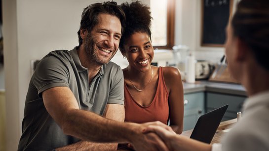 Junges Paar sitzt lächelnd an einem Schreibtisch, er schüttelt die Hand eines Beraters gegenüber