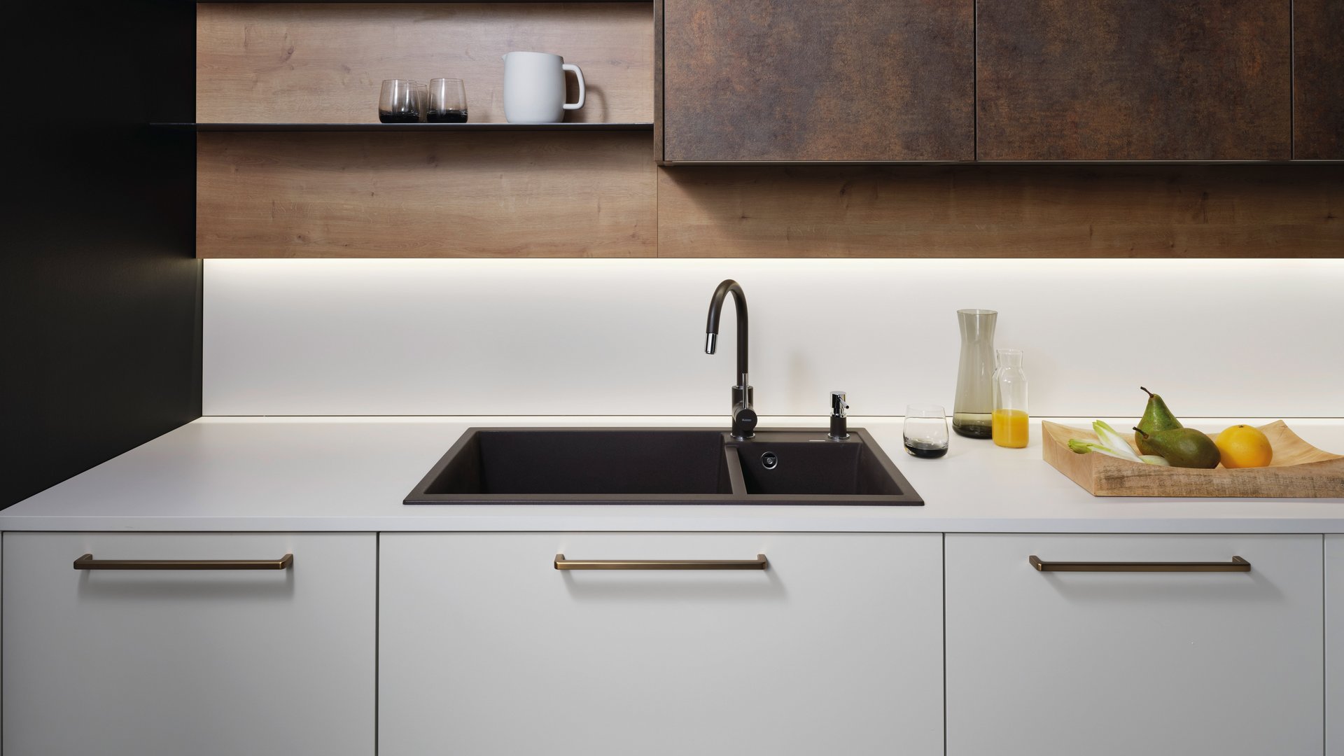 ewe Nuova/Cemento in angora/patina bronze Ausschnitt einer hellen Küchenzeile mit schwarzer Spüle und Armatur, darüber Wandboards, daneben Oberschränke in patina bronze.