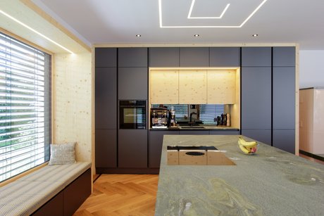 Im Vordergrund eine Kücheninsel mit Arbeitsplatte aus Stein, dahinter eine Küchenzeile in dunkelgrau mit großer holzverkleideter Nische