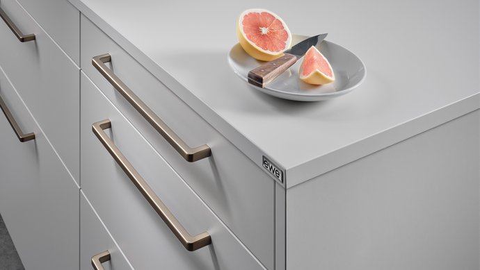 Ausschnitt einer hellgrauen Kücheninsel mit bronzefarbenen Griffbügeln, darauf ein Teller mit Grapefruit