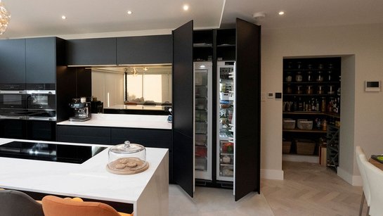 Kücheninsel mit Marmor-Arbeitsplatte, dahinter eine schwarze Küche mit großen, geöffneten Kühlschrank