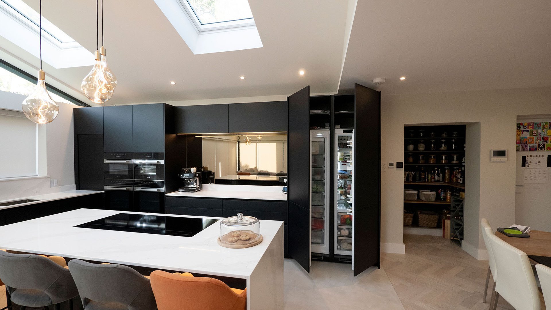 ewe nanoo® in onyxschwarz | Searle & Taylor, UK Kücheninsel mit Marmor-Arbeitsplatte, dahinter eine schwarze Küche mit großen, geöffneten Kühlschrank