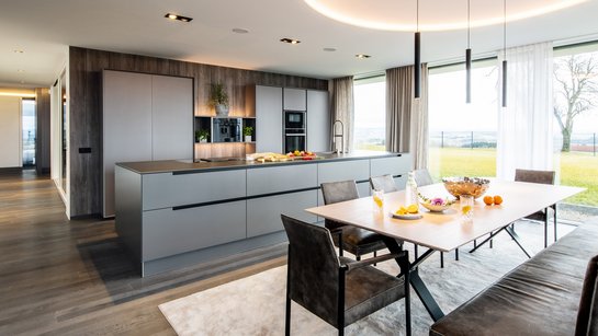 Großer Wohnraum mit Esstischgruppe im Vordergrund, dahinter eine große Inselküche neben einem Panoramafenster