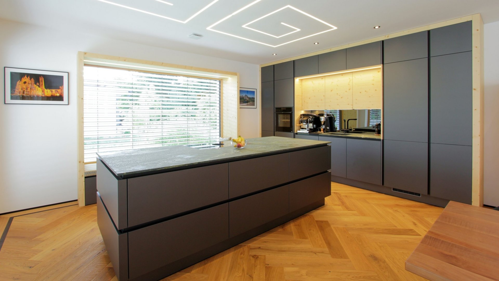 ewe nanoo® in carbon metallic | Laserer Tischlerei & Küchenstudio Große Inselküche in dunkelgrau metallic mit schwarzen Griffmulden mit holzverkleideter Nische in den Hochschränken