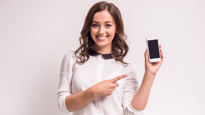 Eine fröhliche junge Frau in einem hellen Pullover zeigt auf ihr Smartphone