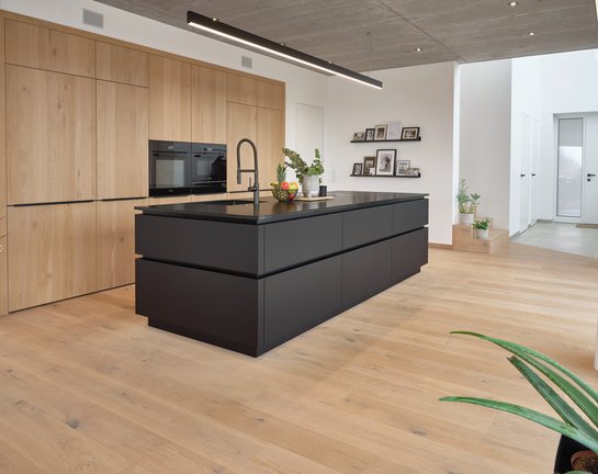 schwarze Kücheninsel mit umlaufender Griffmulde, dahinter deckenhohe Hochschränke mit Holzfronten