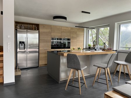 Graue Kücheninsel mit Holz-Thekenplatte und Barstühlen, dahinter eine Hochschrank-Zeile mit Holzfronten