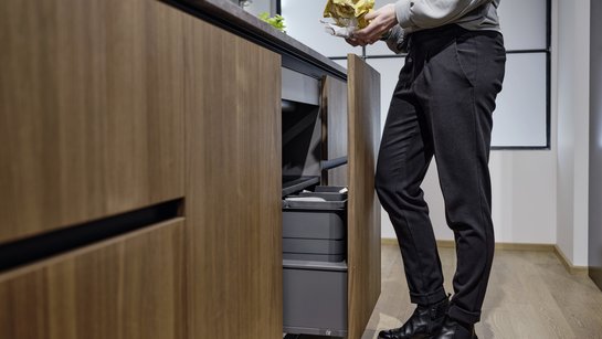 Eine Frau hat Abfall in der Hand und steht in der Küche vor einem Unterschrank mit integriertem Müllsystem, das sie mit dem Knie automatisiert öffnen kann.