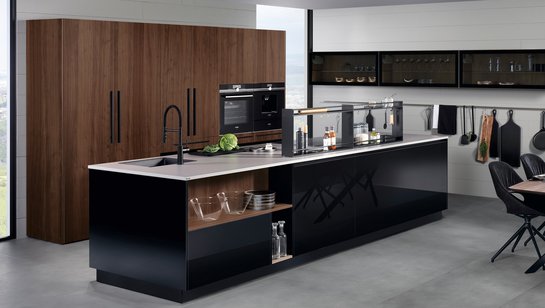 ewe Lucca . ewe Legno in schwarz . ambernuss, schräge Hauptansicht Kücheninsel in schwarz Hochglanz mit Hochschrankgruppe mit Fronten in ambernuss furniert