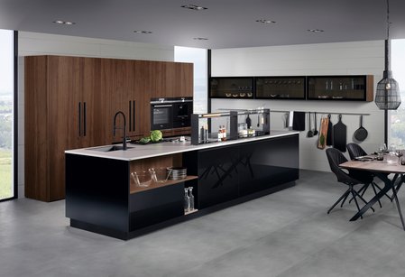 ewe Lucca . ewe Legno in schwarz . ambernuss, schräge Hauptansicht Kücheninsel in schwarz Hochglanz mit Hochschrankgruppe mit Fronten in ambernuss furniert