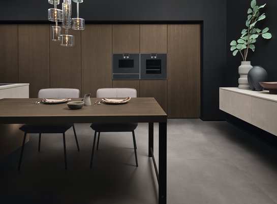 Esstisch in dunklem Holz, dahinter Küchenhochschränke und ein graues Sideboard