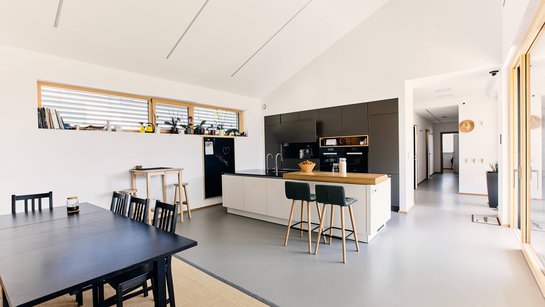 Wohn-Esszimmer mit weiß-grauer Inselküche, davor ein schwarzer Esstisch mit Holzstühlen