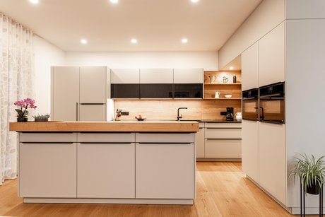 Hellgraue Inselküche mit Holz-Barplatte, Küchenzeile dahinter und Hochschränken rechts