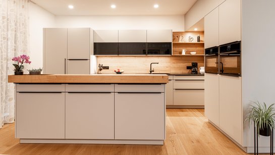 Hellgraue Inselküche mit Holz-Barplatte, Küchenzeile dahinter und Hochschränken rechts