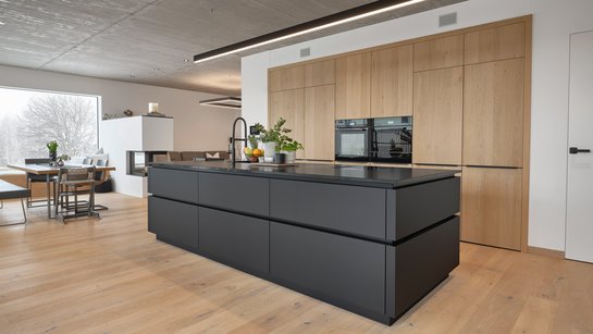 schwarze Kücheninsel mit umlaufender Griffmulde, dahinter deckenhohe Hochschränke mit Holzfronten