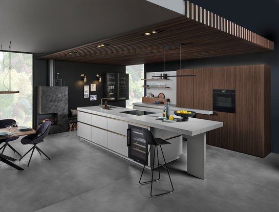 Inselküche in grauer Strukturoberfläche mit bronzefarbigen Griffmulden und einer Hochschrankgruppe in Nuss-Optik