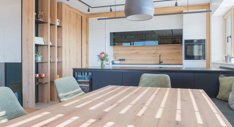 Blick in die Küche mit Raumteiler aus Holzelementen, integrierten offenen Regalen für Deko und einer Küchenzeile in steinweiß mit Holzrückwand und Glasoberschränken.