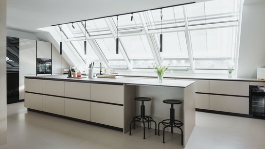 Helle Inselküche mit schwarzer Arbeitsplatte und schwarzen Griffmulden unter schräger Fensterfront