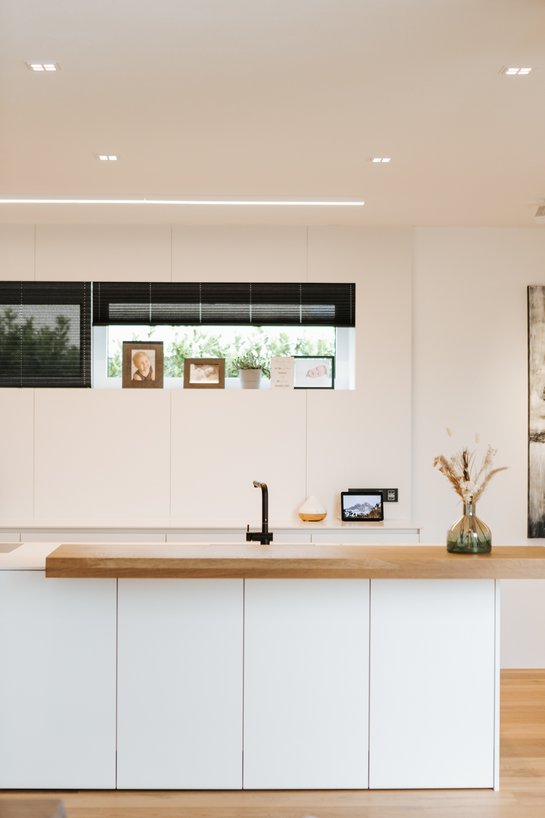 Eine mattweiße Kücheninsel mit einer Barplatte aus Holz, dahinter ein schmales Fenster in der Wand mit Deko devor.
